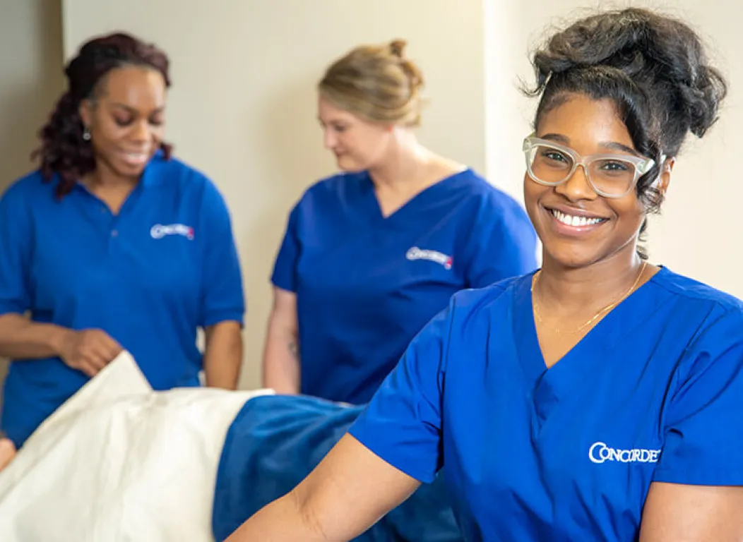 A massage therapist leans on her table and smiles at the camera.
