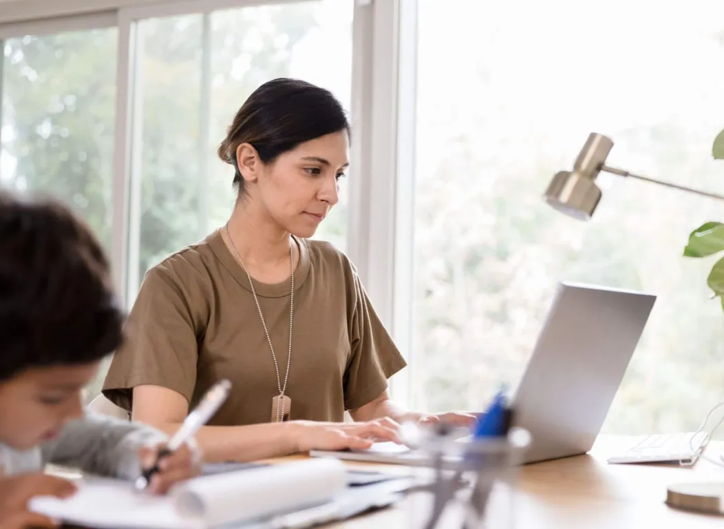 A female veteran with dog tags working on a laptop while her child studies nearby, highlighting the flexibility of Concorde military tuition support for families.