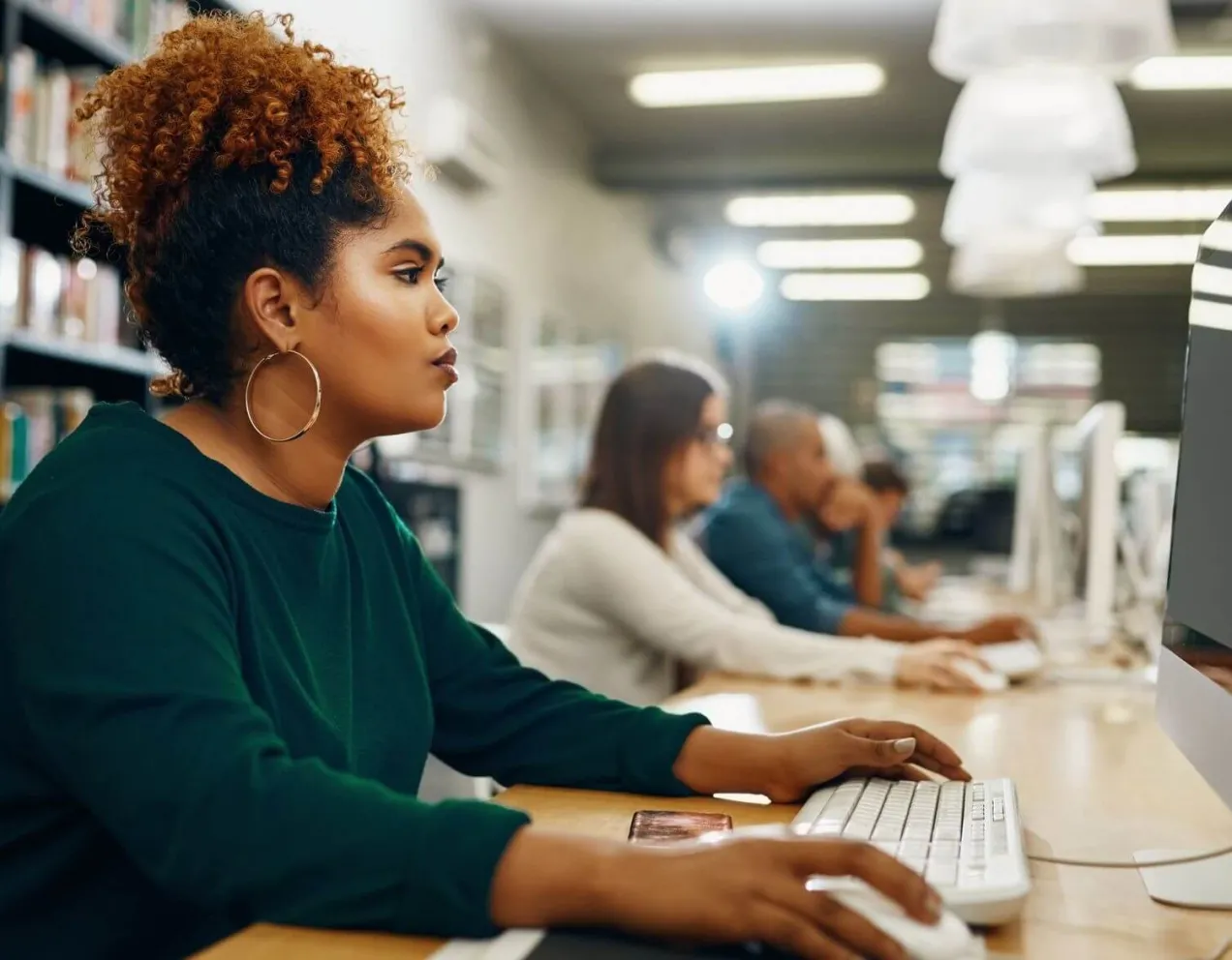 A focused woman with curly hair sits at a computer in a library or lab, working on the coursework that may be part of her healthcare administration degree prerequisites.