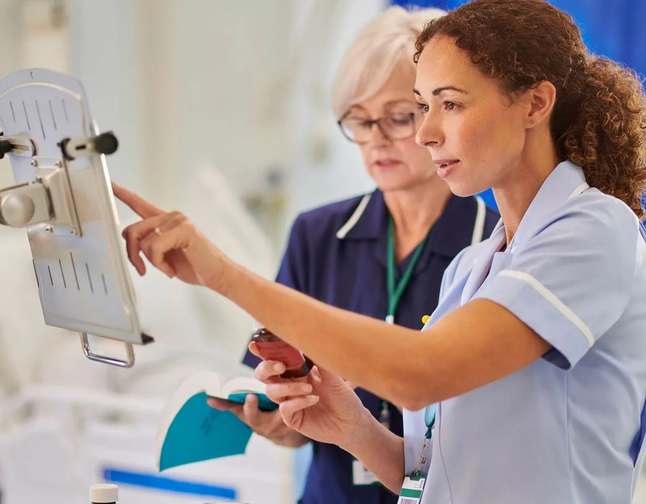 Two female nurses in professional uniforms review medical data on a wall-mounted digital terminal while one holds a medication bottle and a reference book.