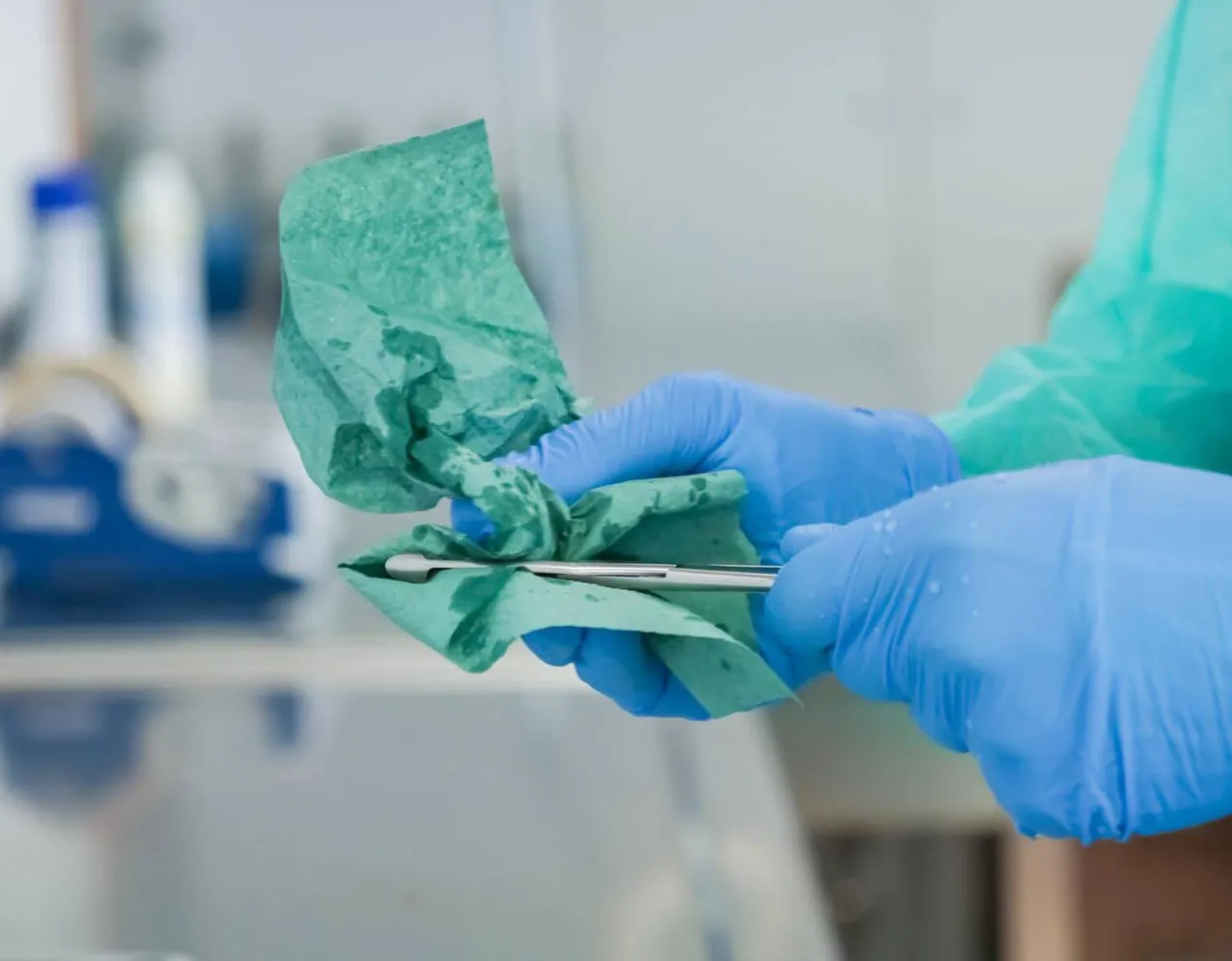 A person wearing a blue protective gown and surgical gloves carefully wipes and cleans a small surgical instrument during the decontamination phase of sterile processing.