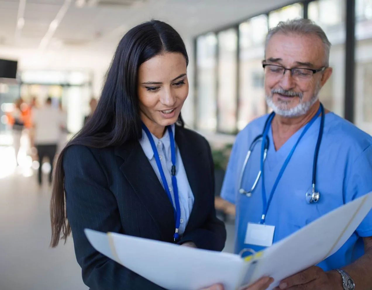 A female healthcare administrator in a black suit reviews medical documents in a binder with a senior male doctor in blue scrubs.