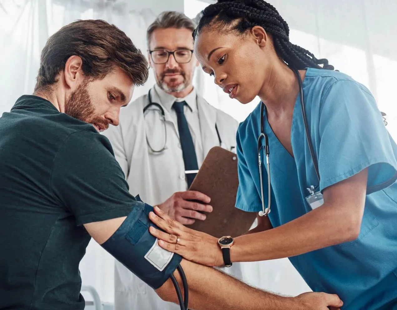 A medical assistant in blue scrubs applies a blood pressure cuff to a patient's arm while being supervised by a senior doctor in a clinical setting.