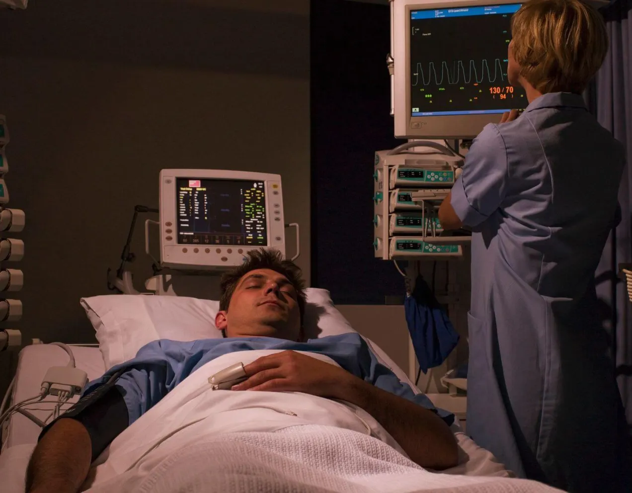 A sleep technologist monitors real-time physiological data and vital signs on multiple screens while a patient sleeps in a darkened room.