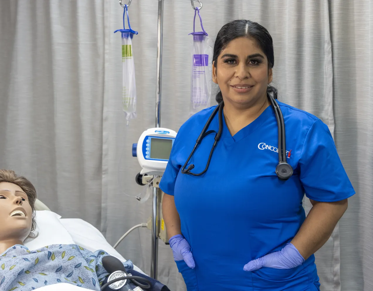 A female nurse with dark hair and blue scrubs poses for camera in lab setting