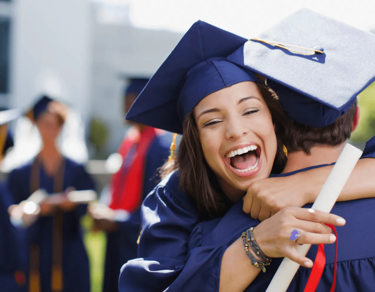 Female graduate in blue cap and gown holding diploma hugs another graduate with big smile on her face.