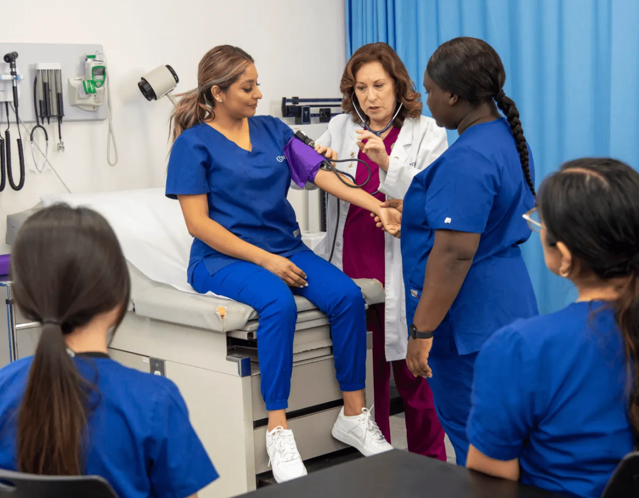 A healthcare professional and educator demonstrates how to take a patient's blood pressure using a student, while other students in the class watch