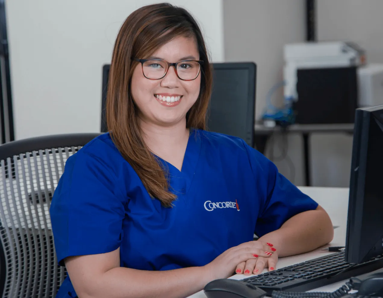 female medical office adminstrator in blue scrubs sits at computer and smiles for camera