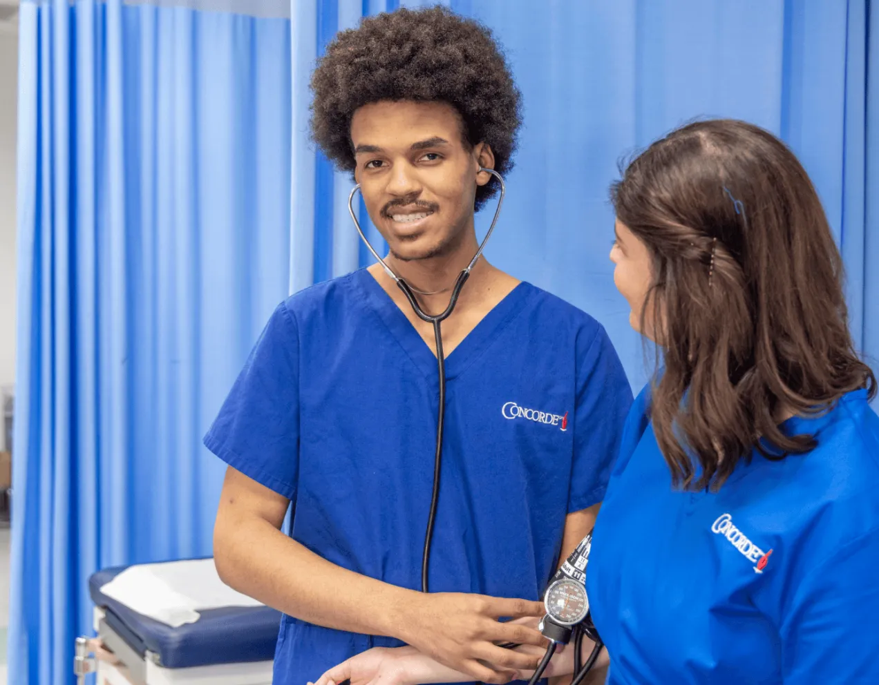 A male medical assistant wearing blue scrubs and stethescope works with patient and smiles for camera