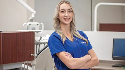 A dental hygienist wearing scrubs in dental office confidently smiles at the camera.