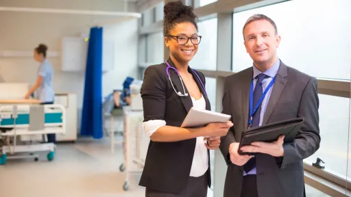 Healthcare administrator and colleague smiling in hospital hallway, holding tablets and reports.