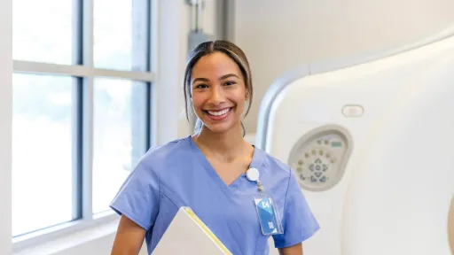 Smiling radiologic technology graduate standing in front of imaging equipment holding medical files.