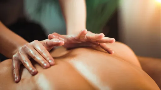 A close-up shot of a therapist's hands massaging the oiled back of a person in a warm, relaxing environment.