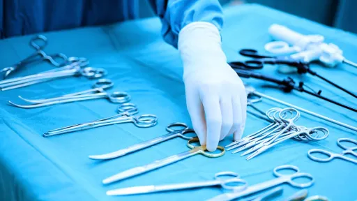 A close-up shot of a gloved hand, belonging to a surgical technician or surgeon, reaching for a pair of surgical scissors among a selection of other medical clamps and instruments on a blue sterile field.