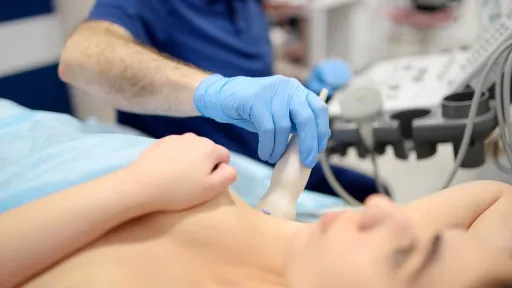 A cardiovascular sonographer in blue gloves performs a cardiovascular ultrasound (sonography) on a woman's chest using a transducer probe.