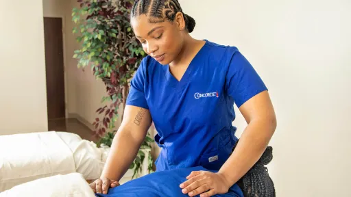 A professional massage therapist wearing blue scrubs with a Concorde logo is performing a leg massage on a client in a bright treatment room.
