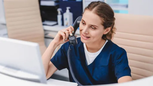 A smiling medical office administrator is seated at a reception desk wearing a blue uniform and smiling while speaking on a landline phone.