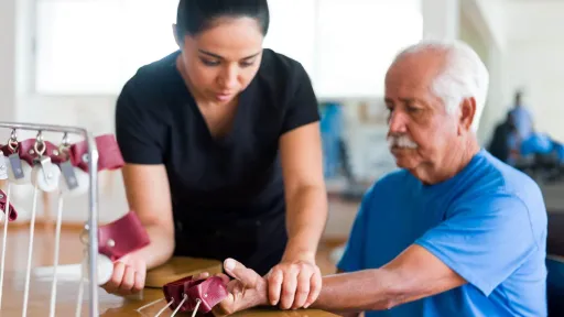Occupational therapy assistant guiding a patient through arm and wrist rehabilitation exercises using therapeutic equipment on a wooden table in a clinical setting.