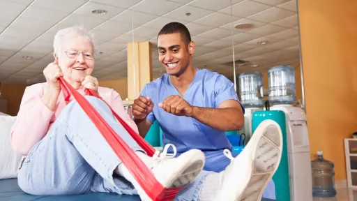Occupational therapy assistant supporting a patient with resistance band leg exercises during a rehabilitation session in a clinical setting.