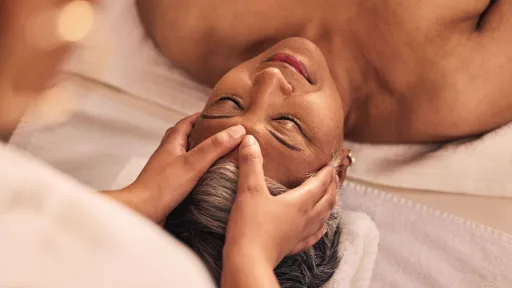 A senior woman with gray hair is lying on a table with her eyes closed while receiving a relaxing head and face massage from a massage therapy professional.