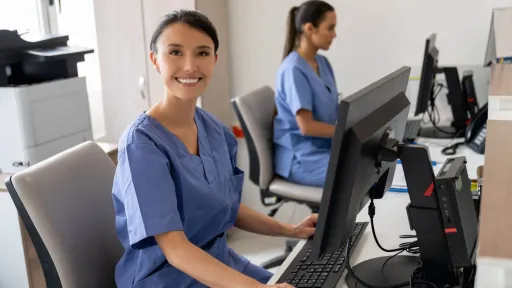 A smiling female medical office administrator in blue scrubs is working at a computer in an office, with a colleague working in the background.