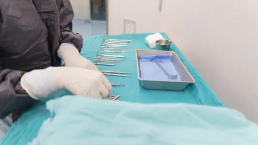 A surgical technologist wearing medical scrubs and gloves organizes a variety of sterile surgical instruments including scissors, clamps, and scalpels on a draped table in an operating room.