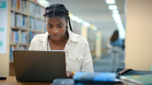 A young Black woman, a student, sits in a library surrounded by bookshelves, concentrating on her laptop while working on her college prerequisites.