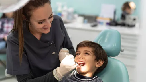 A smiling dental hygienist in a gray uniform and gloves examines the teeth of a young boy in a light-blue dental chair.