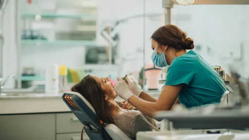 A female dental hygienist wearing scrubs, gloves, and a face mask examines a patient's teeth with attention and detail in a brightly lit dental office.