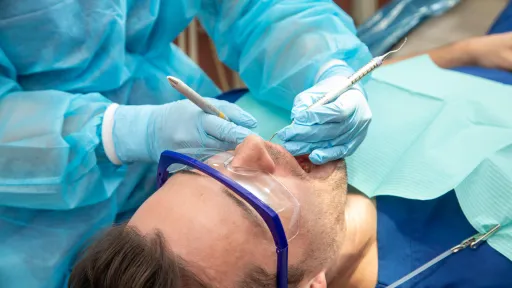 A close-up view of a dental hygienist wearing a blue gown and gloves using dental tools to work on a male patient who is wearing protective eyewear.