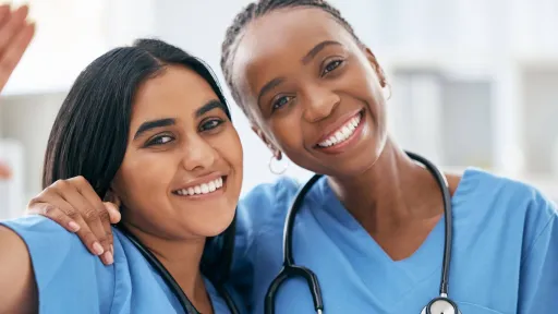 A close-up portrait of two female nurses smiling broadly while standing shoulder-to-shoulder in blue scrubs.