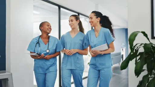 Three diverse female nurses in blue scrubs and stethoscopes smiling and collaborating as they walk down a modern hospital hallway.