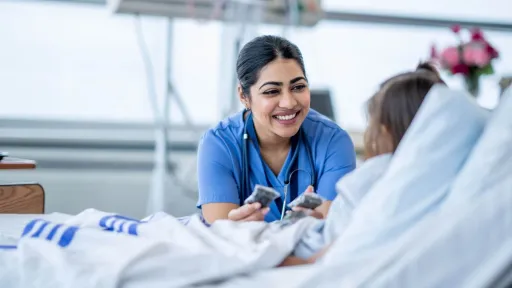 A female nurse in blue scrubs with a stethoscope smiles reassuringly while sitting by a child patient in a hospital bed.