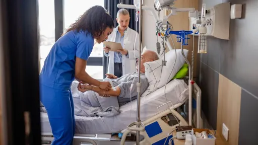 A nurse in blue scrubs provides direct patient care by adjusting an IV line for an elderly man in a hospital bed while a doctor reviews a chart in the background.