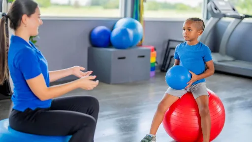 A pediatric physical therapist assistant guides a young child sitting on a specialized balance trainer to improve core stability and stepping coordination.