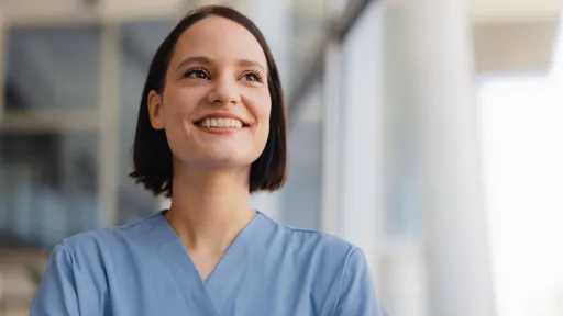A close-up portrait of a smiling female nurse in blue scrubs looking upward with a hopeful expression in a bright, modern healthcare setting.