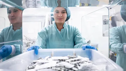 A female sterile processing technician smiles while working in a pharmaceutical cleanroom, wearing protective gear and handling a tray of packaged medication blister packs.