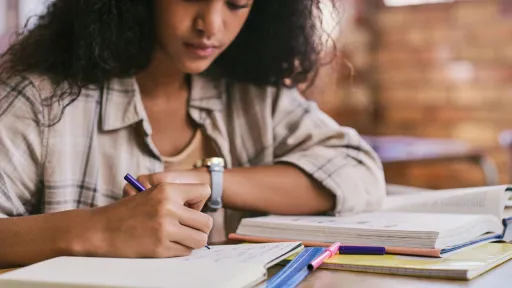 A close-up of a student taking notes and tracking important FAFSA deadlines in a notebook while surrounded by textbooks.