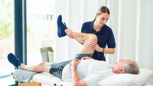 A female physical therapist assistant performs a passive range of motion leg stretch on an elderly male patient in a private clinical setting.
