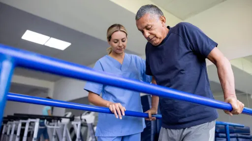 A physical therapist assistant in blue scrubs monitors an older man as he practices walking using parallel bars in a rehabilitation center or hospital gym.