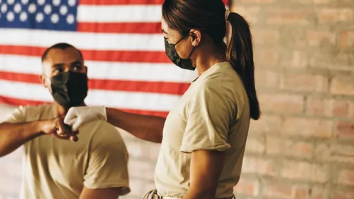 Two military service members wearing face masks and medical gloves give each other a fist bump in front of an American flag, representing the teamwork involved in transitioning from a military medic to an RN.