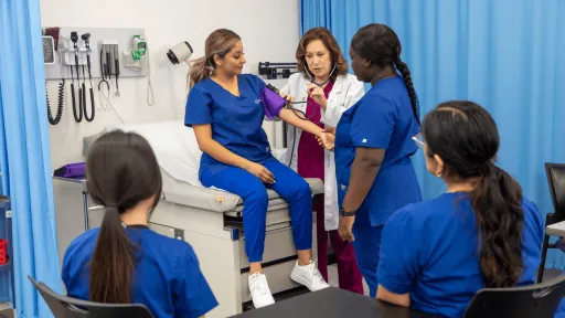 A healthcare professional and educator demonstrates how to take a patient's blood pressure using a student, while other students in the class watch