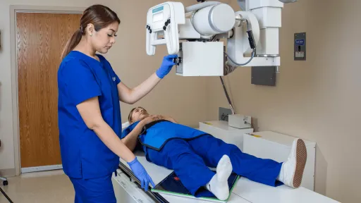 Female radiologic technologist with dark hair and blue scrubs prepares a patient laying on x-ray table for imaging