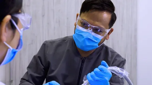 A male dental assistant in grey scrubs and blue face mask holds dental instrument while working with other dental professional