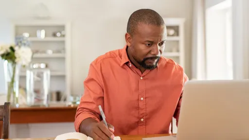Adult man working from home, researching, and studying on a laptop while taking notes.