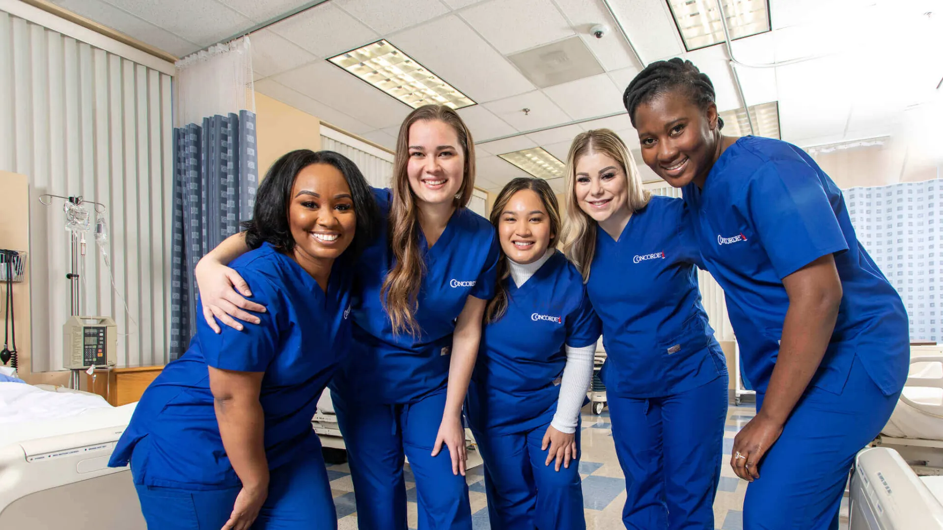 5 smiling Concorde graduates in blue scrubs in lab setting