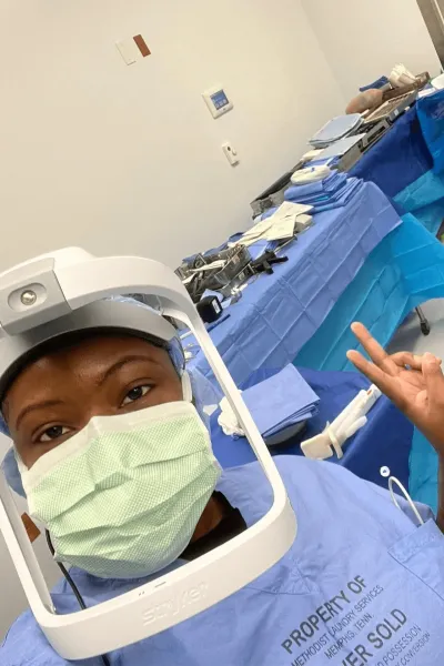 female surgical technologist poses in front of surgical equipment wearing face mask and gear
