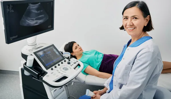 A sonographer smiles at the camera while her patient lies on a bed next to the scanning equipment