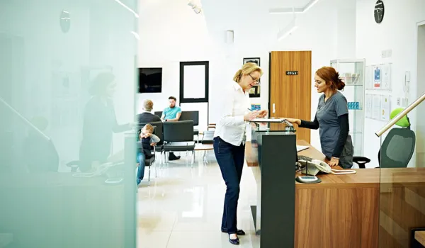 A young medical assistant in scrubs stands behind a desk and interacts with a patient in a waiting room at a medical facility.