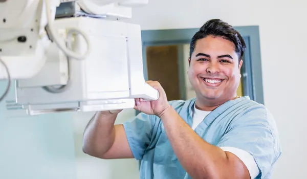 A man wearing scrubs smiles as he positions imaging equipment in a medical facility.